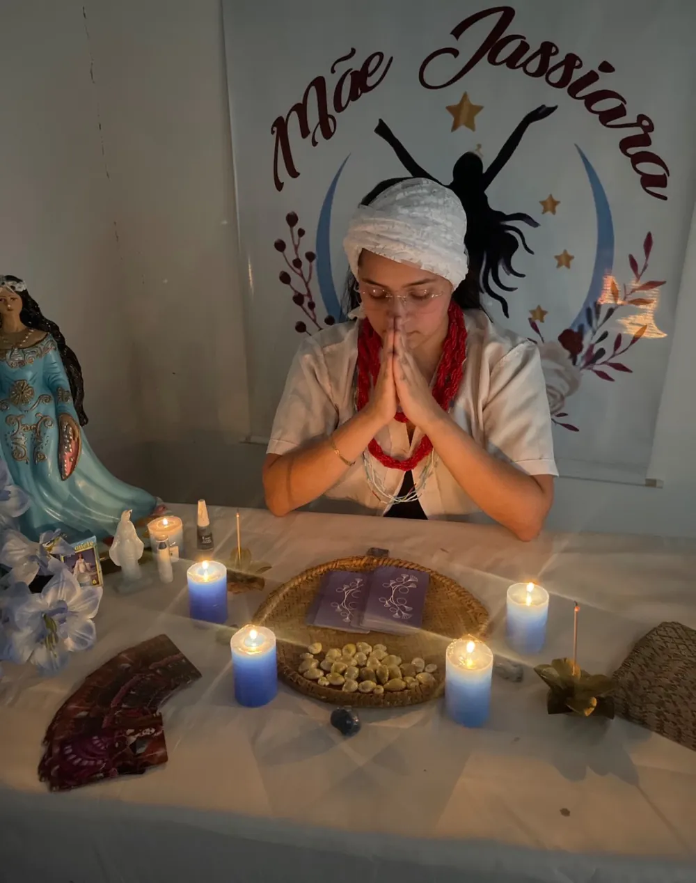 Mãe Jassiara de mãos postas, com turbante branco, diante da mesa de consulta com cartas, búzios e velas azuis