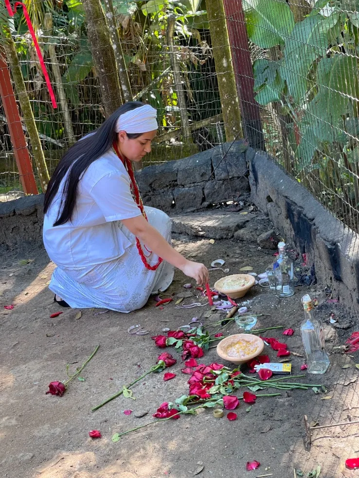 Mãe Jassiara de branco, agachada na mata, arrumando rosas vermelhas e alimentos em uma oferenda