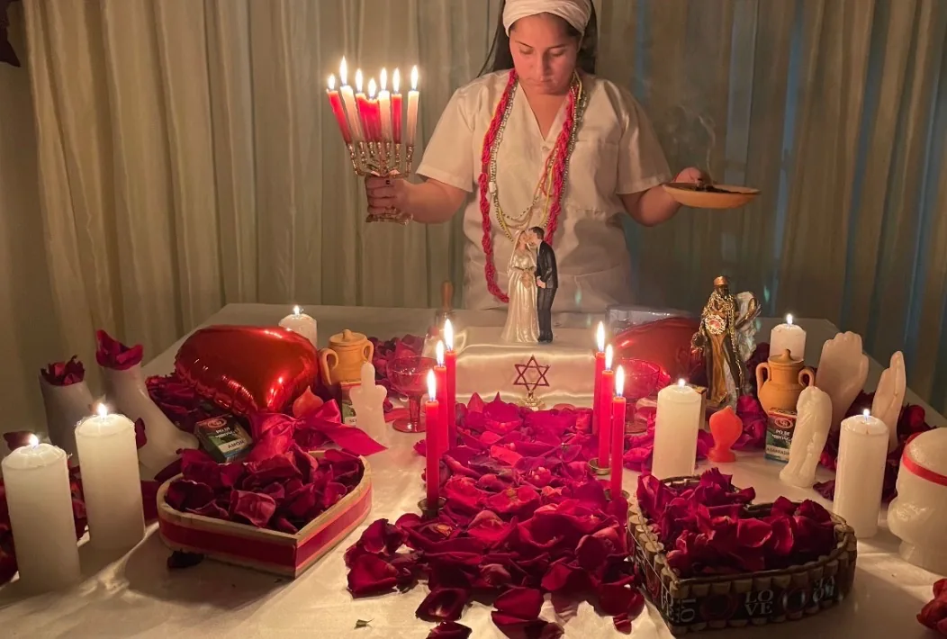 Mãe Jassiara com um candelabro de velas sobre a mesa de união amorosa, coberta de pétalas de rosa e com um casal de noivos